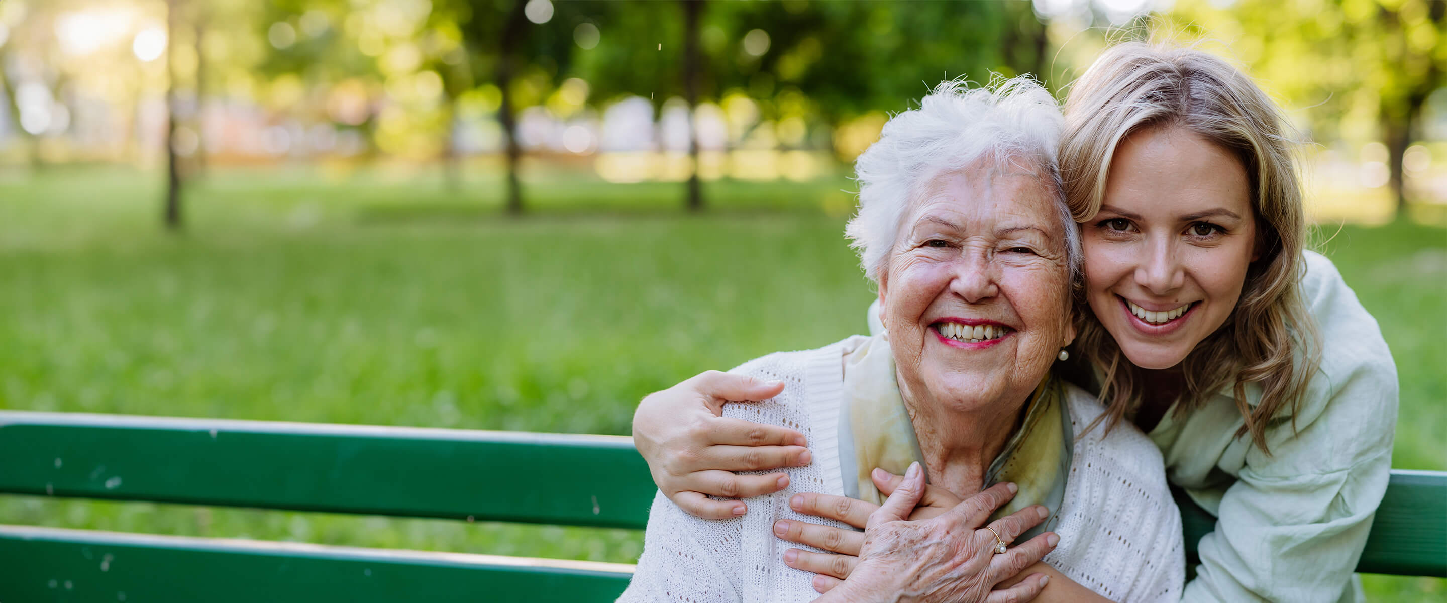 A heartwarming moment between generations: a woman embraces an older woman on a park bench, showing love and care.