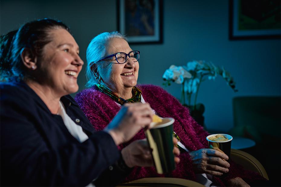 A daughter and mother enjoying drinks and a movie while sitting on a sofa together.