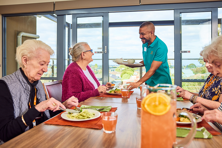 A man kindly serving food to older individuals seated at a table, ensuring their nourishment and care.