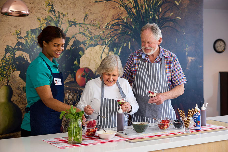 Residents and a staff member participating in a cooking activity at Benetas Corowa Court, preparing and decorating desserts together with a backdrop of a colourful garden mural.