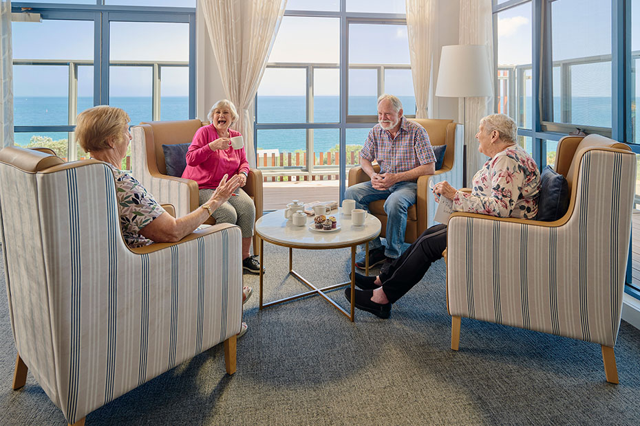 Residents gathering in a relaxing lounge area at Benetas Corowa Court, enjoying tea and conversation with a panoramic view of the bay through large windows.