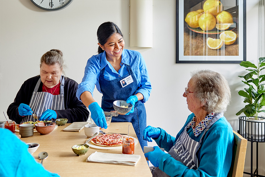 A woman in blue gloves kindly serves food to older individuals, ensuring their nourishment and care.