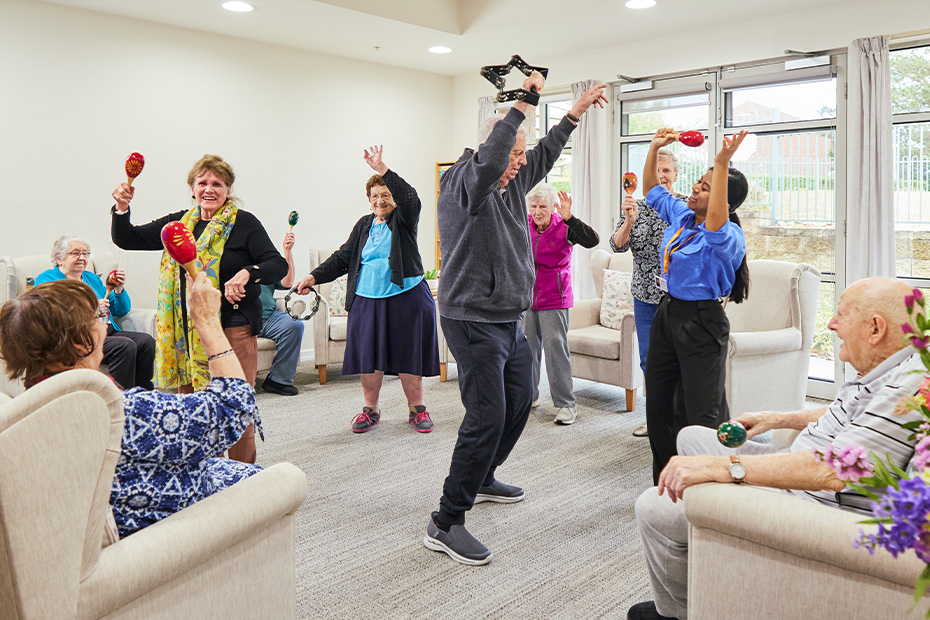 A resident man holding a maraca while a group of people gather in a room.