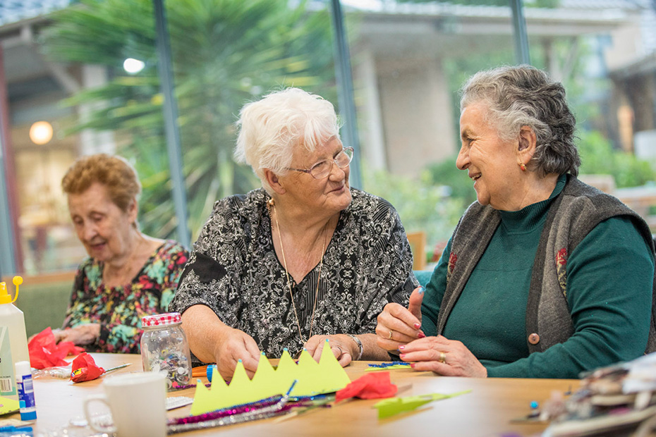 Three older women sitting at a table, engrossed in crafting activities.