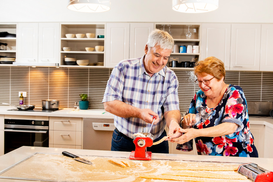 An older couple stands in a kitchen, smiling at each other while preparing a meal together.