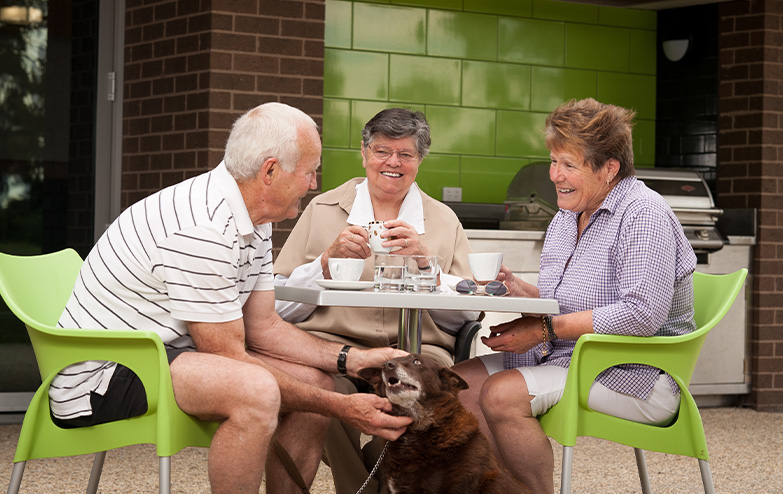 An older couple and their dog enjoying a meal together at an outdoor table.