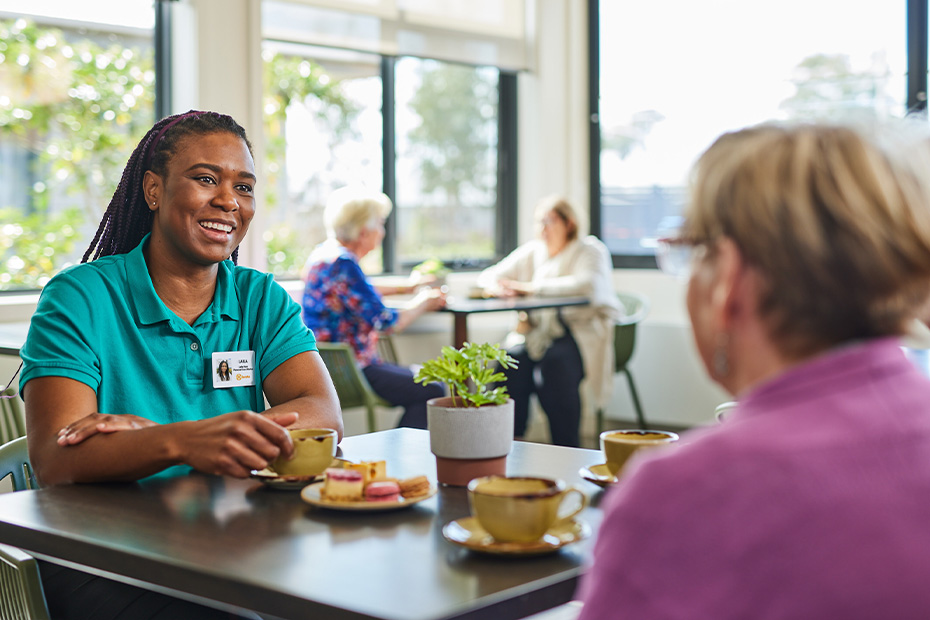 A woman Benetas employee in a green shirt enjoying a cup of tea while sitting at a table with a resident.