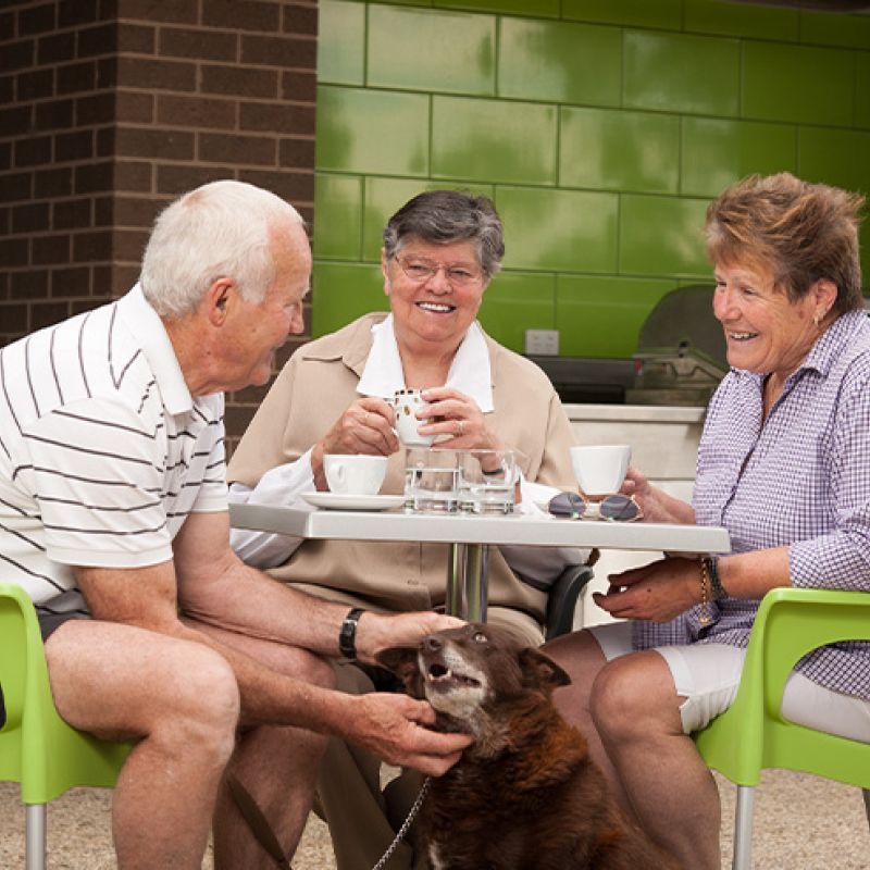An older couple and their dog enjoying a meal together at an outdoor table.